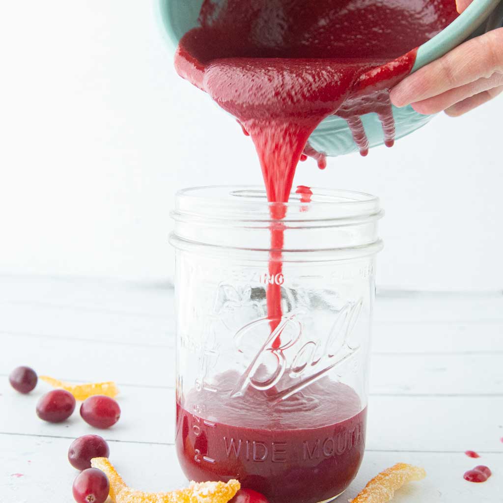 pouring cranberry curd into jar next to whole cranberries and candied orange