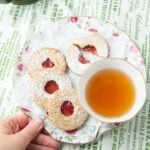 hand holding linzer cookies on plate with cup of tea