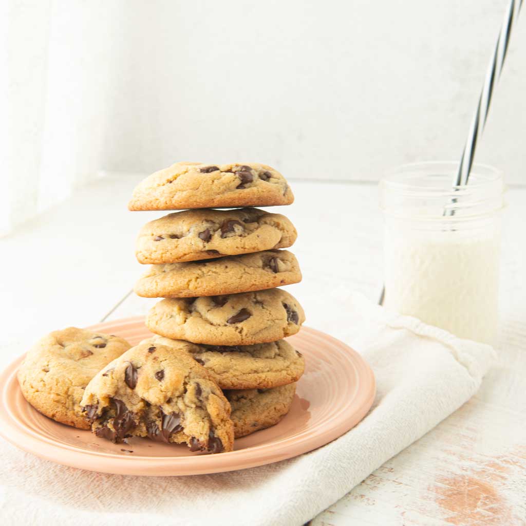 stack of chocolate chip cookies on plate with cup of milk and straw