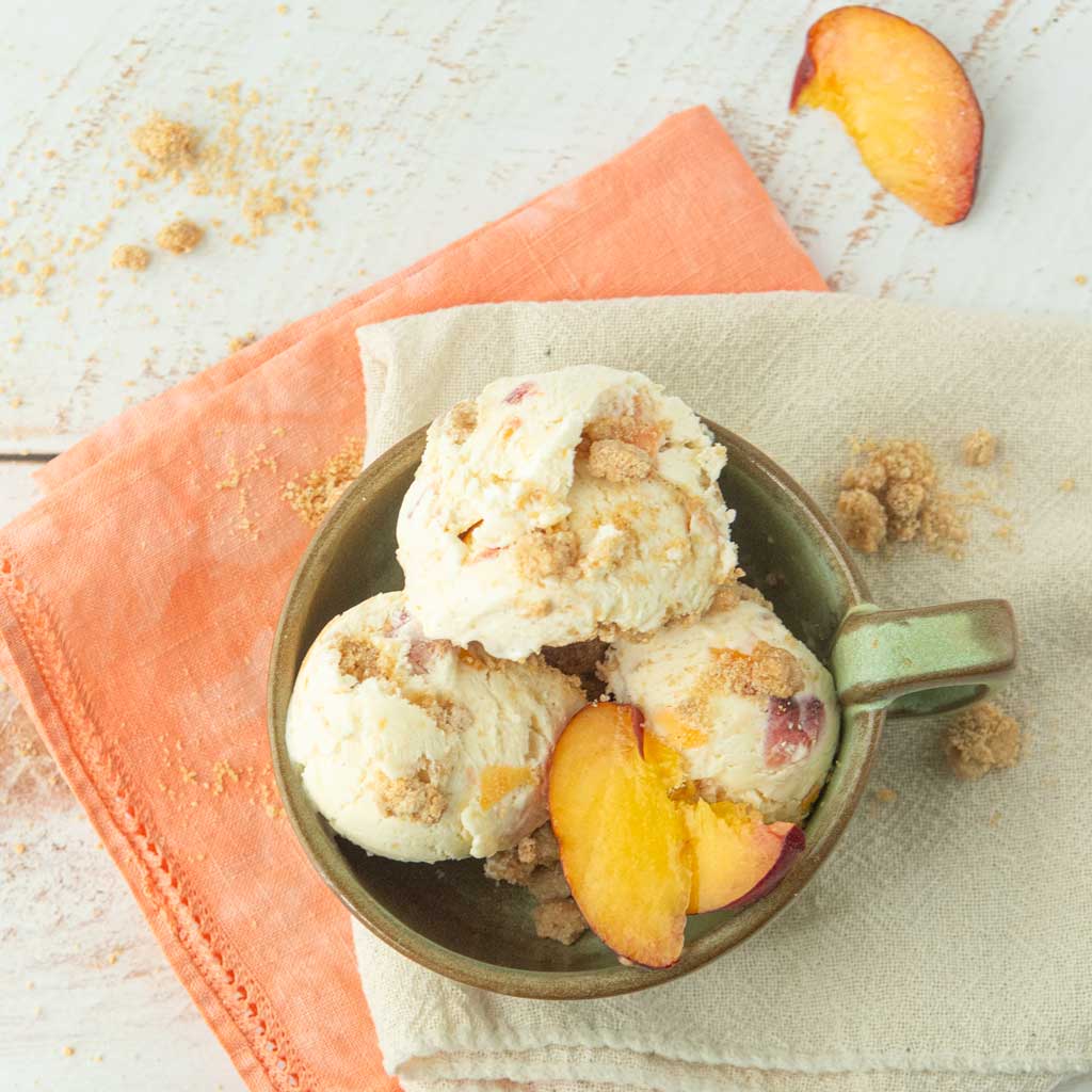 peach ice cream in ceramic dish with peach slices on op of white and peach napkins and extra streusel crumbles