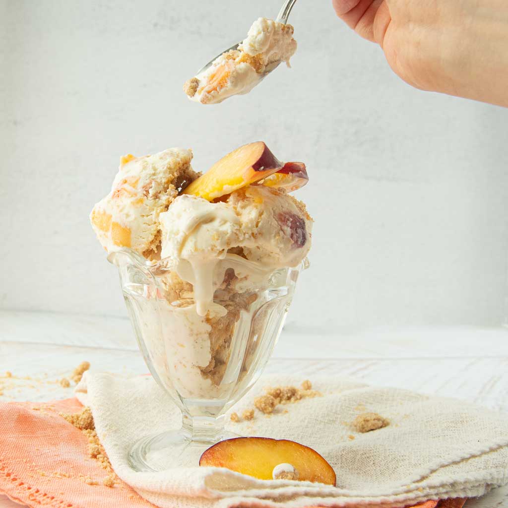 peach ice cream in glass dish with peach slices on op of white and peach napkins with hand showing digging into ice cream