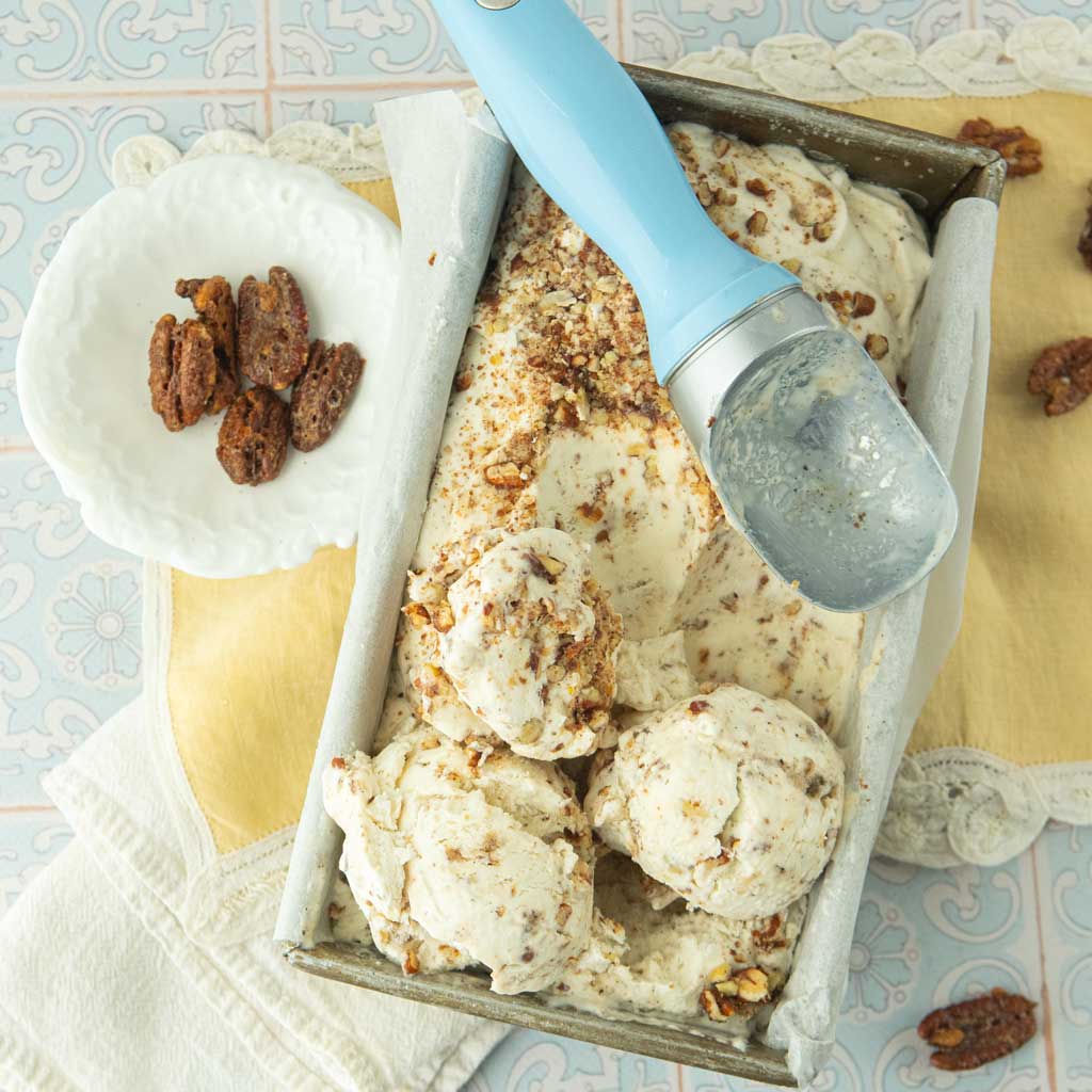 ice cream scoop, dish of candied pecans, loaf pan of buttered pecan ice cream with scoops of ice cream sitting on top