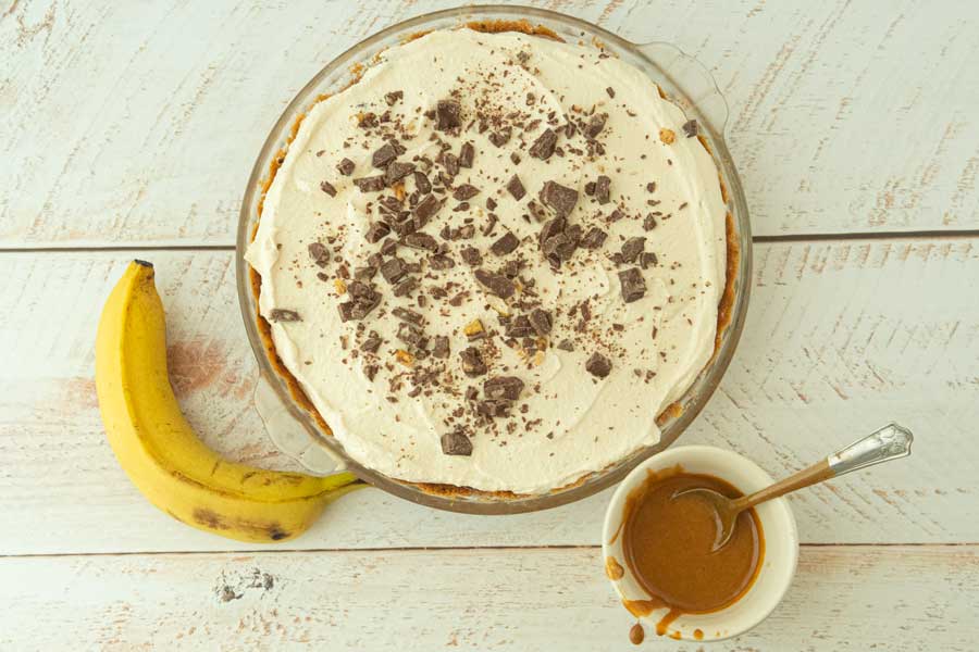 whole banoffee pie covered with toffee candy next to banana and dish of dulce de leche filling