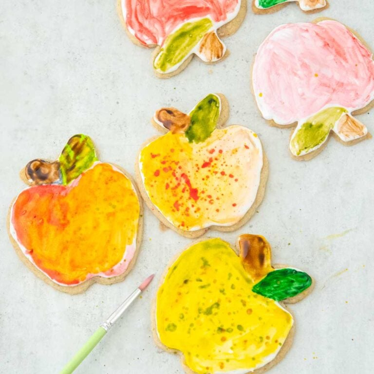 Flat-lay photo of hand-painted apple sugar cookies decorated with colorful edible watercolor icing on parchment paper next to a small paintbrush