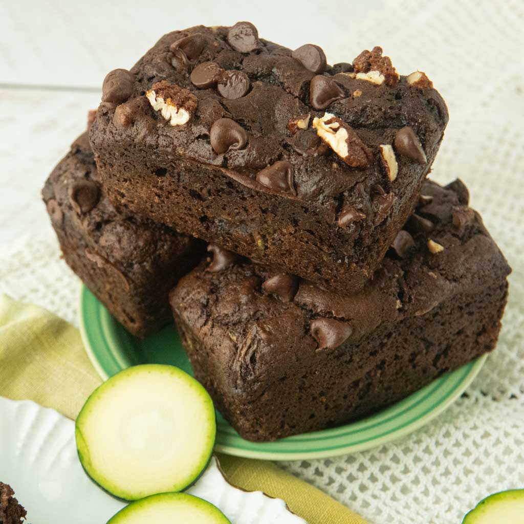 stacked loaves of chocolate zucchini bread on plate next to zucchini slices