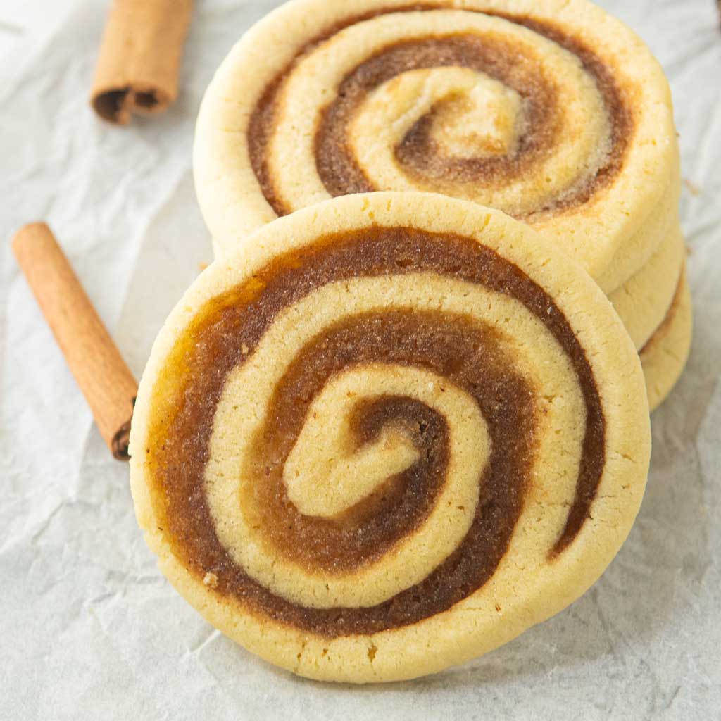 stack of cinnamon roll sugar cookies with one cookie propped against stack, cinnamon sticks