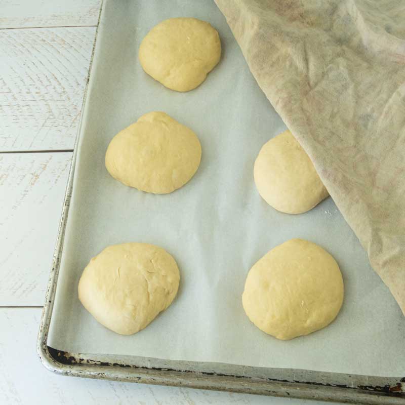 potato buns resting on sheet tray before baking, partially covered by towel